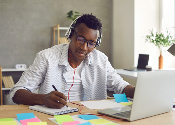 person on computer with headphones taking notes with notepad and sticky notes