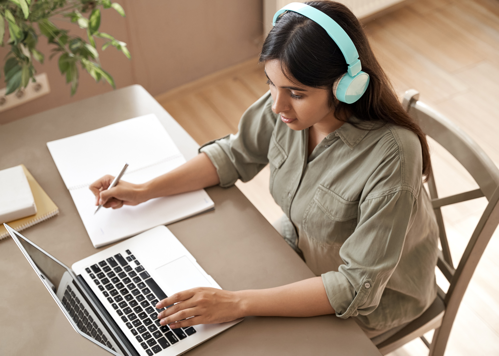 Photo of student wearing headphones working on a laptop and taking notes